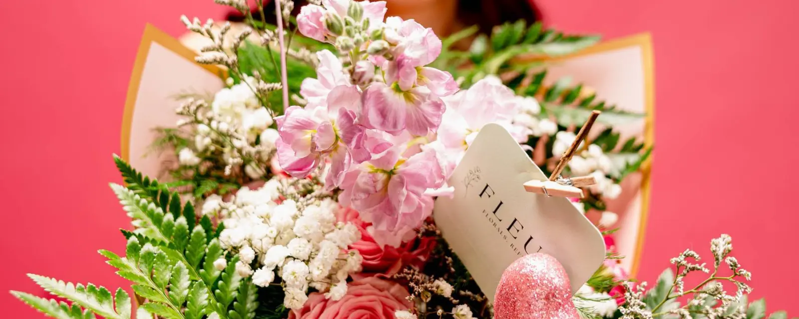 A wrapped bouquet of pink stock, roses and baby’s breath held out toward the camera, a FLEUR. tag clipped to it.