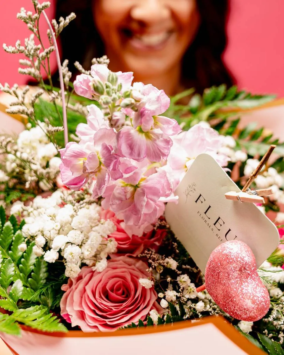 A wrapped bouquet of pink stock, roses and baby’s breath held out toward the camera, a FLEUR. tag clipped to it.